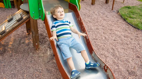 Foto de adorable niño sonriente escalando y montando en la diapositiva. Niño activo divirtiéndose y jugando en el parque — Foto de Stock