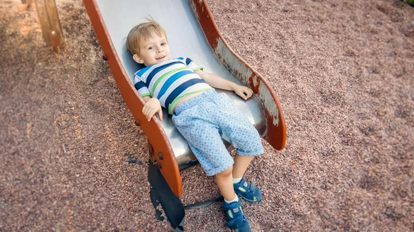 Zdjęcie cute 3 lat chłopiec wspinaczka i jazda na duży slajd na plac zabaw dla dzieci w parku — Zdjęcie stockowe