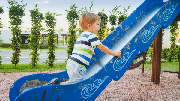 Photo of cute 3 years old toddler boy climbing and riding on big slide on children playground at park — Stock Photo, Image