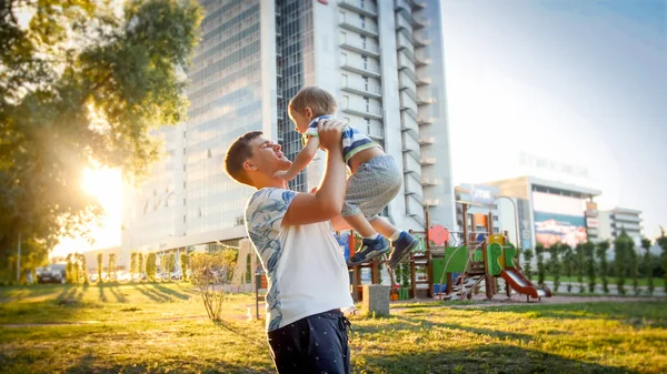Portrait of happy smiling young father holding and throwing up his laughing 3 yearas old little son in park on the susnet — Stock Photo, Image