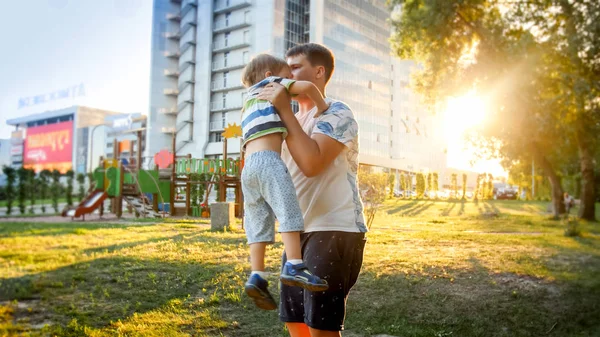 Portrait of happy smiling young father holding and throwing up his laughing 3 yearas old little son in park on the susnet — Stock Photo, Image