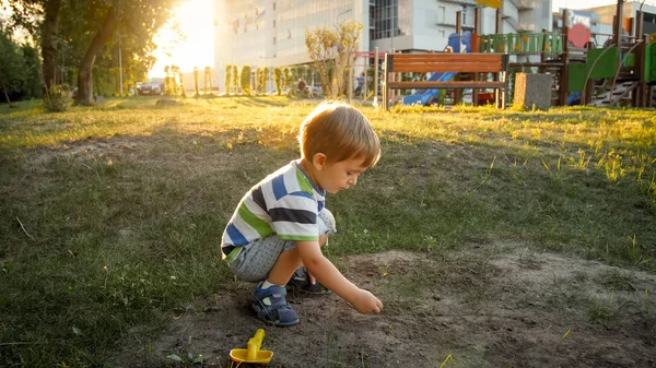 Zdjęcie 3-letniego małego chłopca siedzącego na ziemi w parku i kopania piasku — Zdjęcie stockowe