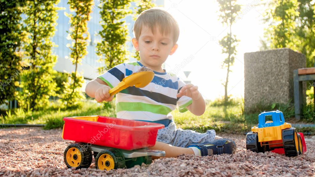 Portrait of adorable 3 years old toddler boy playing with toy truck with trailer on the playground at park. Child digging and building from sand