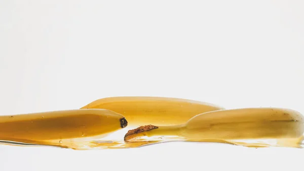 Foto de cerca de frutas frescas maduras que caen en el agua. Babanas amarillas dulces salpicando y fluyendo en agua sobre fondo blanco —  Fotos de Stock