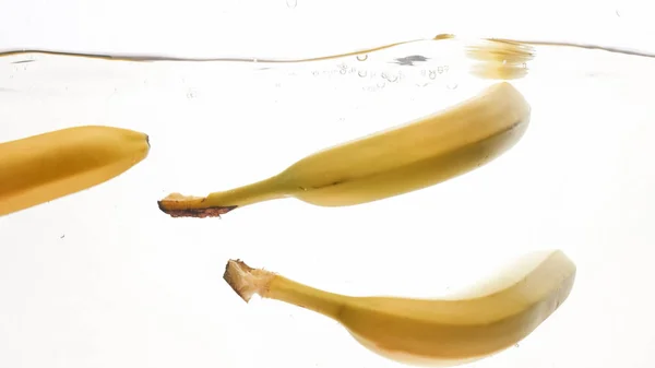 Φωτογραφικά πλάνα από φρέσκες ώριμες κίτρινες μπανάνες που πέφτουν και πιτσιλίζουν σε καθαρό νερό από απομονωμένο λευκό πίσω — Φωτογραφία Αρχείου