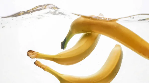 Κοντινό πλάνο φωτογραφίες φρέσκων φρούτων που πέφτουν στο νερό. Κίτρινο γλυκό babanas πιτσίλισμα και ρέει στο νερό από λευκό φόντο — Φωτογραφία Αρχείου