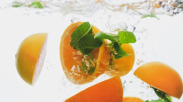 Imagen de cerca de naranjas jugosas frescas cortadas con hojas de menta flotando en agua clara sobre fondo blanco — Foto de Stock