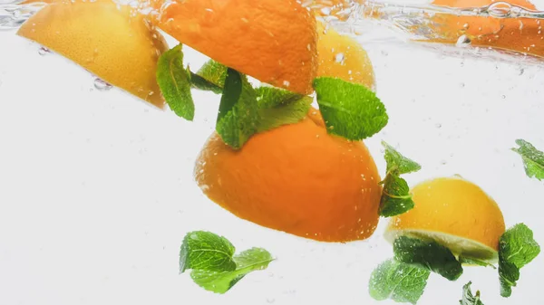 Imagen de cerca de naranja jugosa fresca cortada y un montón de hojas de menta cayendo y salpicando en el agua clara contra el fondo blanco — Foto de Stock
