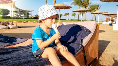 Komik portre sevimli gülümseyen bebek çocuk güneş yatağında oturan ve dondurma yalama. Yaz plaj tatil tatilde rahatlatıcı çocuk