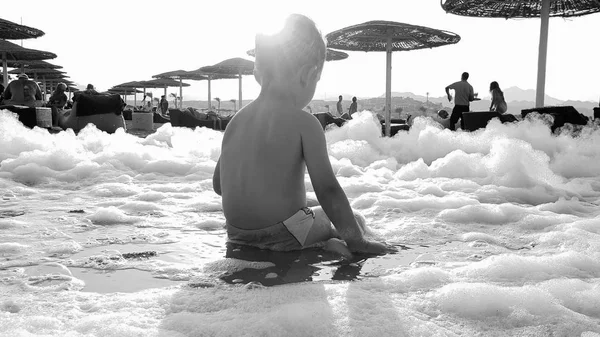 Foto de primer plano de un niño pequeño de 3 años sentado en la playa de arena y jugando con mucha espuma de jabón después de la fiesta disco de playa — Foto de Stock