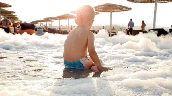 Imagen tonificada del adorable niño jugando con espuma de jabón en la playa de arena de mar —  Fotos de Stock