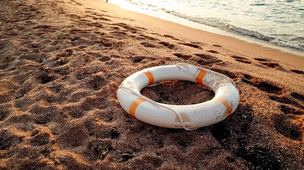 Imagen de primer plano del anillo salvavidas de plástico blanco que se encuentra en la playa de arena contra la hermosa puesta de sol sobre el agua —  Fotos de Stock