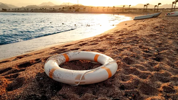 Closeup image of white plastic life saving ring lying on the sandy sea beach against beutiful sunset over the water — Stock Photo, Image