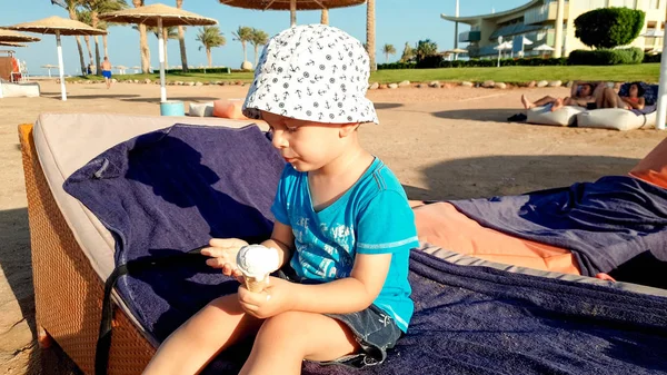 Zabawny portret ofcute uśmiechnięty chłopiec maluch siedzi na solarium i lizanie lody. Dziecko relaks na letnich wakacji wakacje na plaży — Zdjęcie stockowe