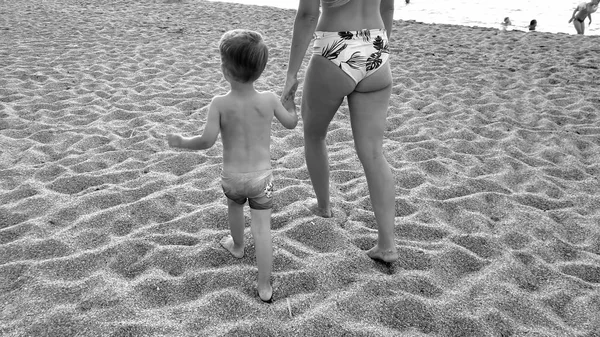 Imagen en blanco y negro de la joven madre sosteniendo a su pequeño hijo a mano y caminando por la playa de arena — Foto de Stock