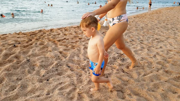 Imagen de la feliz madre joven con adorable niño de 3 años de edad, cogido de la mano y caminando en la playa del mar contra el cielo hermoso atardecer sobre la superficie del agua —  Fotos de Stock