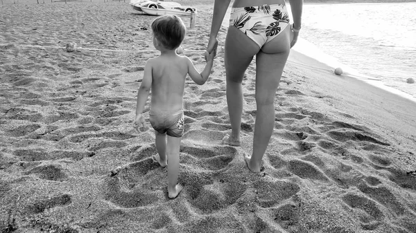 Zwart-wit beeld van jonge moeder houdt haar kleine zoon met de hand en wandelen op het zandstrand van de zee — Stockfoto