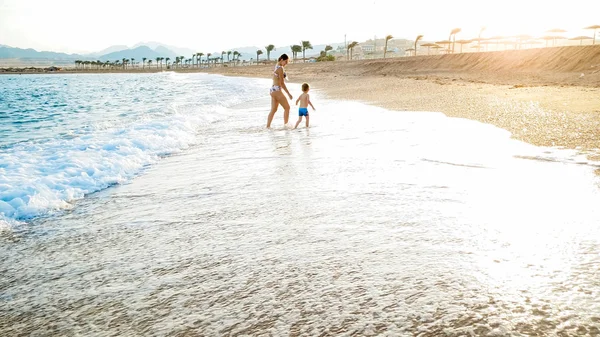 Hermosa foto de la feliz madre joven sosteniendo a su pequeño niño a mano y caminando en la playa de mar sndy en olas cálidas y tranquilas contra el increíble cielo puesta de sol —  Fotos de Stock