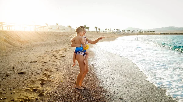 Gyönyörű kép a fiatal anya gazdaság és átölelve neki 3 éves kisfiát a homokos tenger parton, és keresi a nyugodt tenger hullámai és naplemente ég — Stock Fotó
