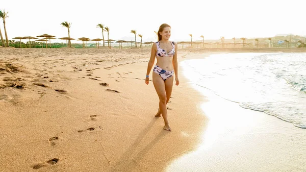 美丽的年轻女子在比基尼的肖像与完美的身体走在平静的海浪对惊人的日落天空 — 图库照片