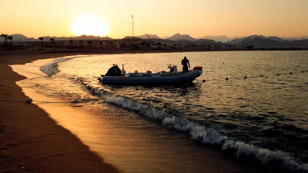 Sorprendente silueta foto de barco inflable con mecedora a motor en las olas del océano calma en la orilla arenosa contra el cielo puesta de sol — Foto de Stock