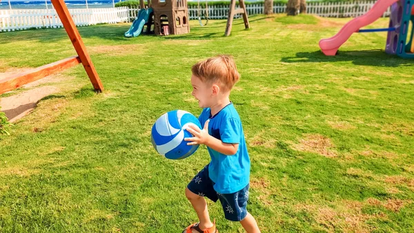 Porträtt av glad leende lille pojke som håller bollen i händerna och springer på gräs på barnens lekplats — Stockfoto