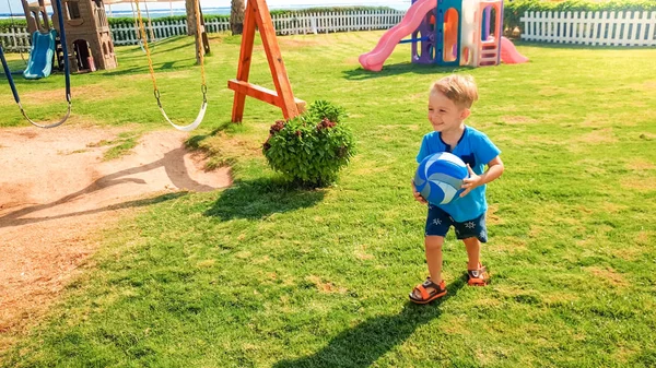 Image of happy laughing cheerful boy holding football ball in hands and running on children playground at park — Stock Photo, Image