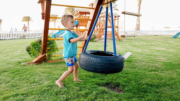 Zdjęcie adorable uśmiechnięty mały chłopiec gra z chaing huśtawka na placu zabaw. Aktywne dzieci bawią się i mają na zewnątrz w parku — Zdjęcie stockowe