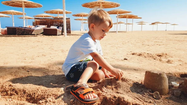 Portret cute 3 lat chłopiec chłopca siedzi na piaszczystej plaży i gry z zabawek i budynku zamek z piasku — Zdjęcie stockowe
