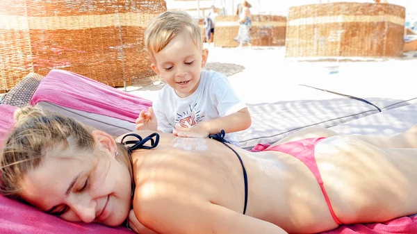 Foto van kleine peuter jongen aanbrengen zonnebrandcrème op moeders terug liggend op ligstoel op zee strand — Stockfoto