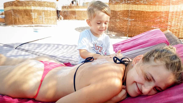 Portret van jonge moeder liggend op de zonnebank op het strand, terwijl haar kind zoon maken van haar een massage — Stockfoto