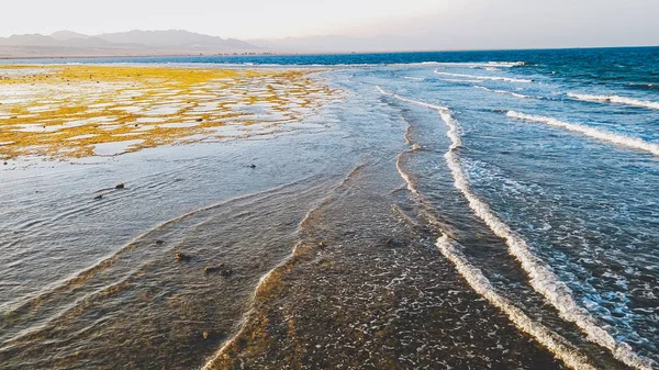 Όμορφο τοπίο με ήρεμο ωκεανό και τα βουνά στην ακτή. Κύματα θαλάσσης κυλώντας και φρενάρετε πάνω από νεκρούς κοραλλιογενείς υφάλους και βράχους στο φως του ηλιοβασιλέματος — Φωτογραφία Αρχείου