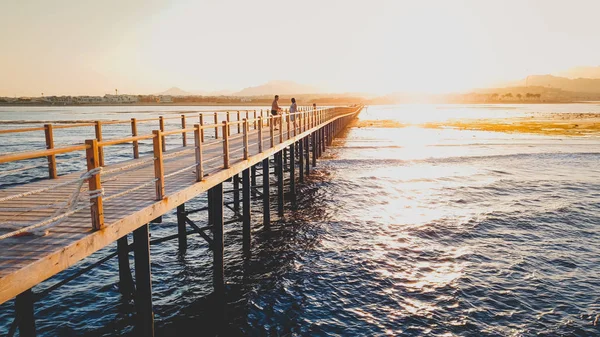 Gyönyörű táj a nyugodt óceán hullámai és a hosszú, fából készült mólón. Csodálatos móló a tengeren ellen naplemente a hegyek felett — Stock Fotó