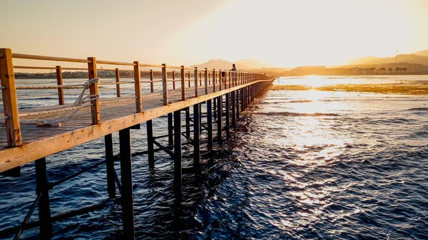 Beautiful landscape of calm ocean waves and long wooden pier. Amazing jetty at sea aagainst sunset over the mountains — Stock Photo, Image