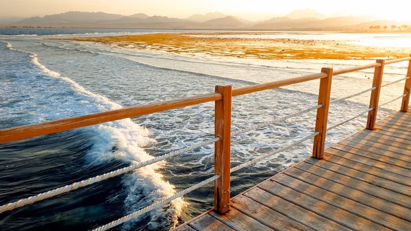 Hermoso paisaje de tranquilas olas oceánicas y largo muelle de madera. Increíble embarcadero en el mar contra el atardecer sobre las montañas —  Fotos de Stock