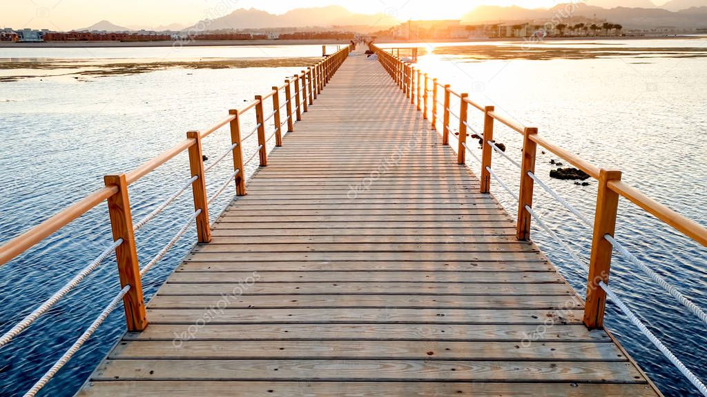Beautiful landscape of calm ocean waves and long wooden pier. Amazing jetty at sea aagainst sunset over the mountains