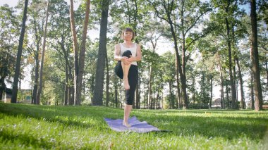 Orta yaşlı gülümseyen mutlu kadın meditasyon ve ormanda çim üzerinde yoga egzersizleri yapıyor Görüntü. Parkta fitness ve germe pracitising ise onun fiziksel ve ruhsal sağlığı bakımı Kadın