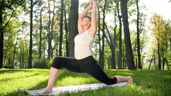 Imagem de meados da idade sorrindo mulher feliz meditando e fazendo exercícios de ioga na grama na floresta. Mulher cuidando de sua saúde física e mental enquanto pratica fitness e alongamento no parque — Fotografia de Stock