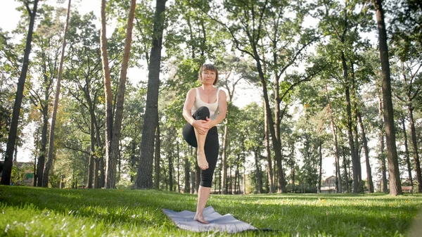 Photo of middle aged woman in sportrs clothes practicing yoga outdoors at park. Middle aged woman stretching and meditating at forest — Stock Photo, Image