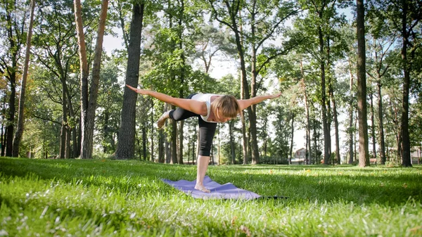 Wizerunek uśmiechniętej kobiety w średnim wieku w fitness ubrania robi rozciąganie i ćwiczenia jogi. Kobieta medytacja i czyny sportowe na mat fitness na trawie w parku — Zdjęcie stockowe