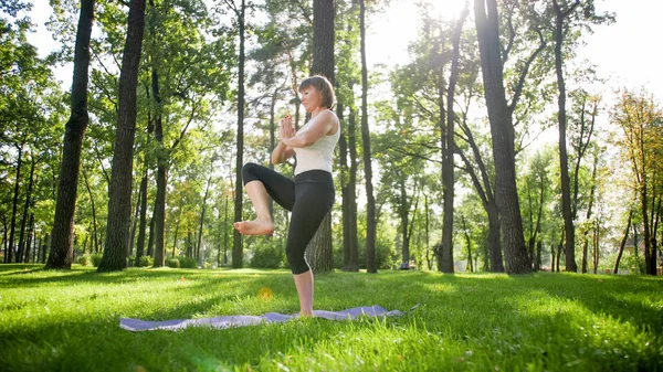 Photo of smiling happy woman 40 years old doing yoga exercises on fitness mat at forest. Harmony of human in nature. Middle aged people taking car of mental and physical health