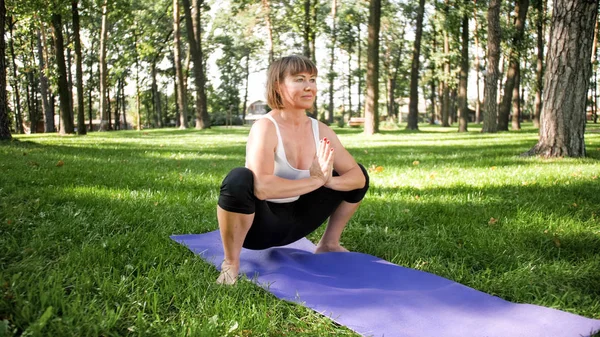 Фото улыбающейся женщины средних лет, практикующей йогу и медитирующей в парке. Женщина растягивается и занимается фитнесом на коврике в лесу — стоковое фото