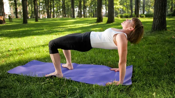 Photo of smiling happy woman 40 years old doing yoga exercises on fitness mat at forest. Harmony of human in nature. Middle aged people taking car of mental and physical health