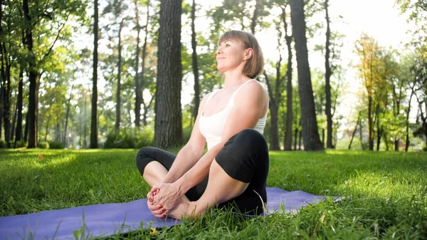 Imagem de meados da idade sorrindo mulher feliz meditando e fazendo exercícios de ioga na grama na floresta. Mulher cuidando de sua saúde física e mental enquanto pratica fitness e alongamento no parque — Fotografia de Stock