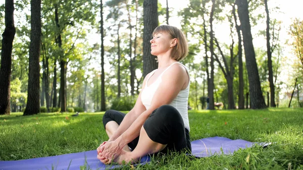 Photo of middle aged woman practicing yoga or fitness on fresh green grass at park. Female physical and mental health. Person in meditation and harmony pf body and soul