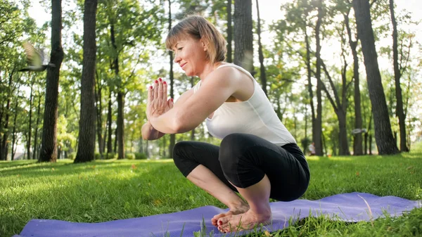Imagem de mulher de meia-idade sorridente em roupas de fitness fazendo alongamento e exercícios de ioga. WOman meditando e fazendo esportes no tapete de fitness na grama no parque — Fotografia de Stock