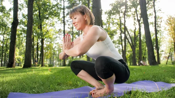 Imagen de mujer feliz sonriente de mediana edad meditando y haciendo ejercicios de yoga sobre hierba en el bosque. Mujer cuidando de su salud física y mental mientras practica fitness y estiramiento en el parque — Foto de Stock