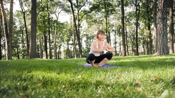 Foto de mulher sorrindo de meia-idade praticando ioga e meditando no parque. Mulher se alongando e fazendo fitness no tapete na floresta — Fotografia de Stock