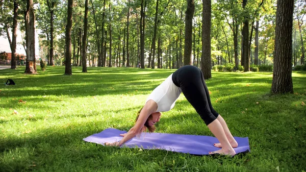 Foto einer lächelnden, glücklichen Frau, 40 Jahre alt, die Yoga-Übungen auf einer Fitnessmatte im Wald macht. Harmonie des Menschen in der Natur. Menschen mittleren Alters, die ein Auto mit geistiger und körperlicher Gesundheit nehmen Stockbild