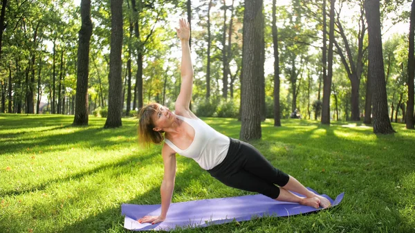 Foto einer lächelnden, glücklichen Frau, 40 Jahre alt, die Yoga-Übungen auf einer Fitnessmatte im Wald macht. Harmonie des Menschen in der Natur. Menschen mittleren Alters, die ein Auto mit geistiger und körperlicher Gesundheit nehmen lizenzfreie Stockfotos